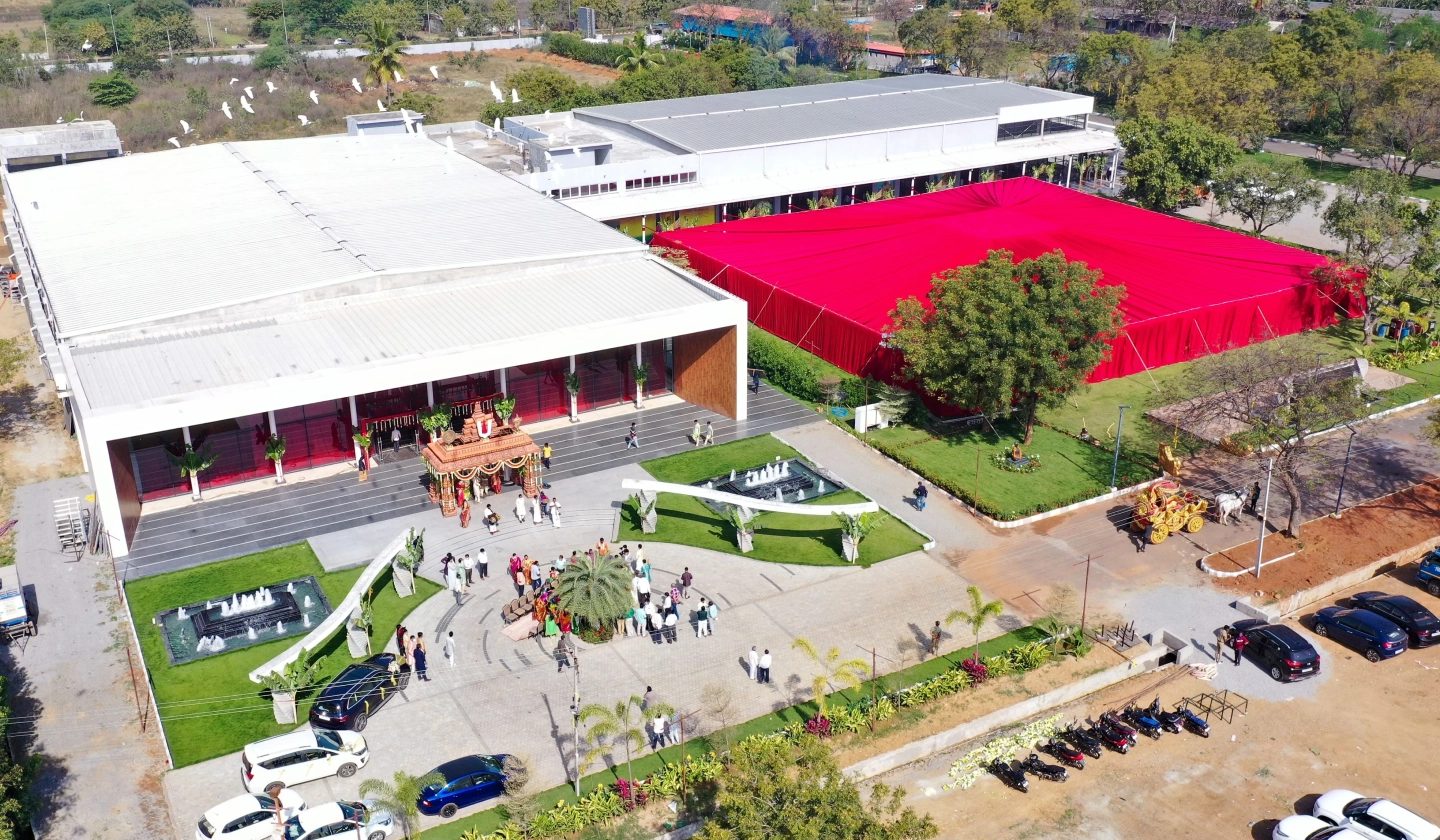 Aerial photograph during an event: a red shamiana pitched beside the hall block, guests on the paved forecourt beside the fountains, cars and two-wheelers parked along the edge.
