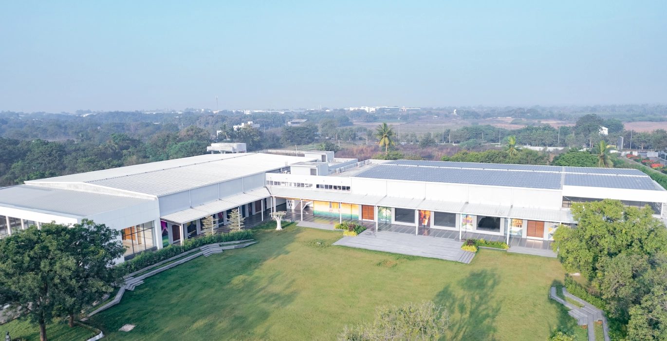 Aerial photograph of the property: the long white hall block, the open lawn along its garden side, the tree belt at the boundary and Madikonda in the distance.